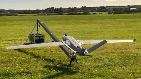 BBC A Tekever AR3 drone, which is shaped like a plane with large wings and a tail, in a field