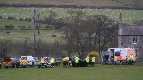 PA Media Members of a search and rescue team arrive at River Aln near Alnwick, Northumberland. There are four vehicles and people wearing hi-vis jackets standing by them, hills can be seen in the background.