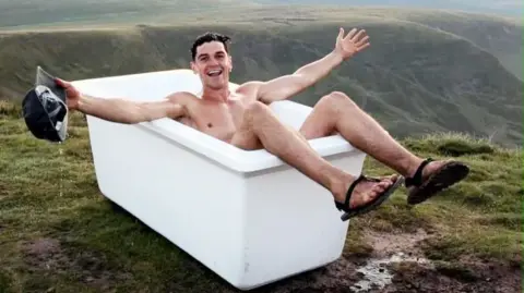 A man is bathing in a bathtub posing for a camera while on a mountain summit. He has short dark hair, holds a black cap in one hand, and is wearing flip flops. 