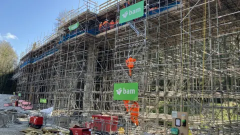 BBC The image shows a lot of building and construction work going on, with lots of scaffolding. There are three workers dressed in orange hi-vis clothing climbing the scaffolding. There are also generators and fencing on the ground. 