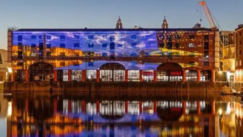Adam Kenrick Huge projected images are reflected onto a brick building on the water. The images reflect back into the water in the evening's darkness. The projected pictures show blue rippling water.
