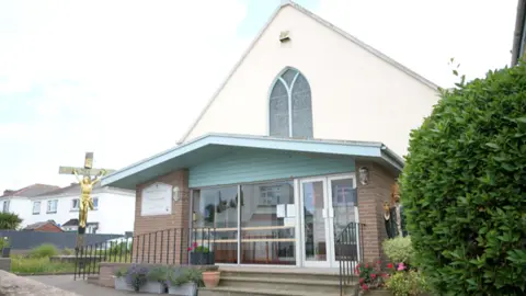 A large cross with a gold coloured image of Christ is outside a church with a brick porch area and a white sloping upper storey. A large stained glass window is in the middle of the wall.