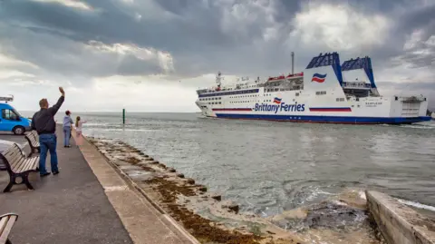 Getty Images Brittany Ferries' Barfleur ferry passing close to shore with three people standing waving on a footpath close to the sea