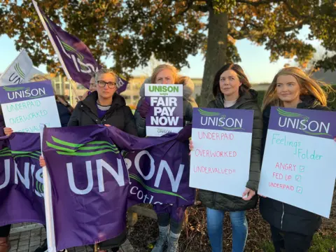 Four women hold Unison placards on the picket line outside a school in Perth. It is a bright autumn day and they are standing in front of a tree with the sun glinting on the yellow-tinged leaves