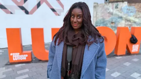 Danny Fullbrook/BBC A woman dressed inn a blue coat with long dark hair is stood in front of giant orange letters spelling the word "Luton"