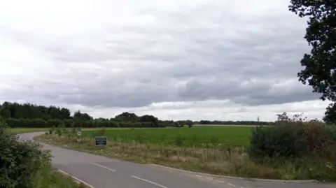Google A narrow road is shown winding through a rural landscape of green crops, hedgerows and trees under leaden skies.