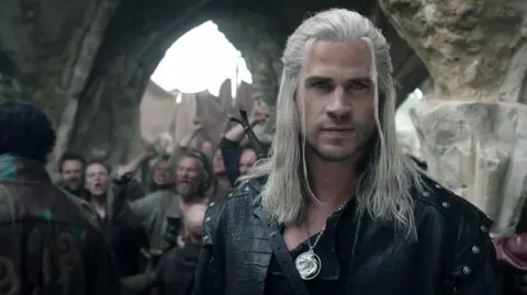 Netflix Liam Hemsworth como Geralt com armadura de couro preto e uma espada no coldre nas costas. Ele tem um pingente de prata e longos cabelos brancos. Ele olha para longe. Atrás dele está um grupo de pessoas furiosas em uma área de estilo medieval.