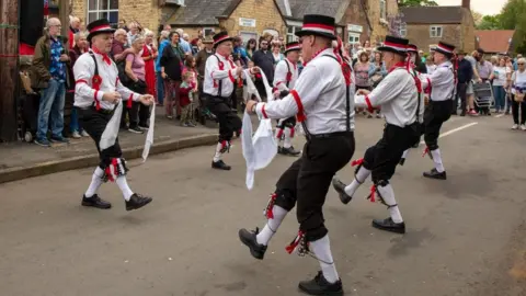Electric Egg Morris dancers dressed in white shirts, black trousers and black shoes with black, red and white hats are mid-dance in the middle of a village street. Spectators can be seen around them.