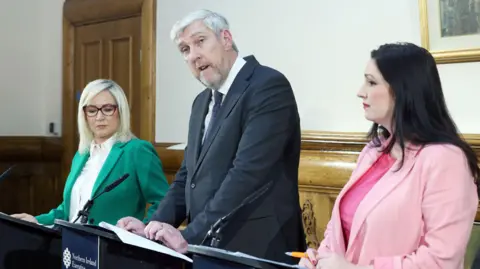 Pacemaker Michelle, John and Emma are standing at podiums in front of a white wall and brown door. 
Michelle is wearing a white shirt and green blazer, with red glasses and blonde short hair.
John is wearing a navy suit with a dark blue tie with pink spots on it.
Emma is wearing a baby pink suit. She has long brown hair and red lipstick on. 