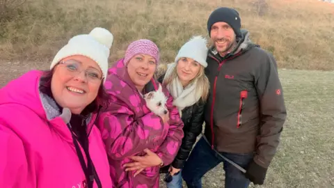 Three women and a man standing in a row on dry grass in warm jackets and woolly hats. Two of the women are in bright pink jackets with a small white dog tucked into one of them. The other woman is in a black leather jacket and white scarf and the man is in a brown waterproof. They are all smiling.