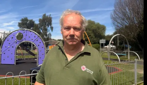 BBC / Jasmine Lowe The photo shows a man looking at the camera. He's wearing a green polo shirt. He's standing in front of a play park. There is a purple climbing wall behind his left shoulder, and a swing behind his right shoulder. It is also fenced off with silver railings. 