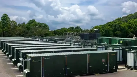 Statera  A row of about 20 green energy storage units about the size of a lorry container with trees around the site 