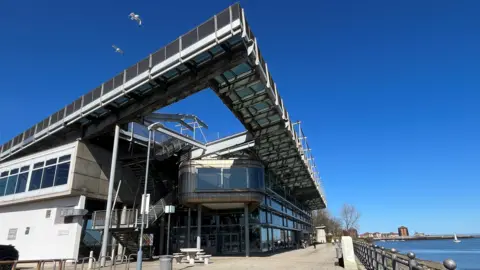 The multy-storey Naional Glass centre building is mostly made of glass and grey panels. It is by the river Wear and has a seating area in front of it. Two seagulls are flying in the clear blue sky above.