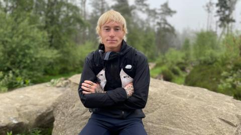 A blonde haired man sits on a rock with folder arms