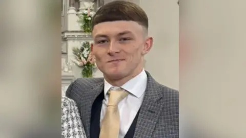 A man in a grey chequered suit, white shirt and gold tie looks at the camera. Behind him are flowers on the wall. 