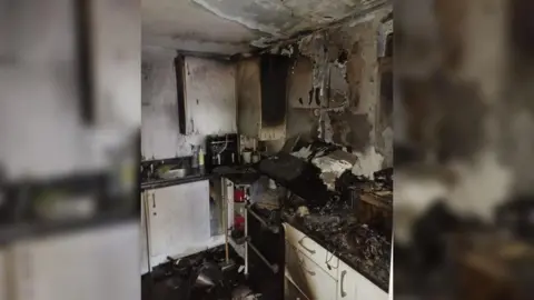 HIWFRS A kitchen which has melted worktops and is covered in black soot. The cooker hood has collapsed and fallen off the wall and on to the hob below. The white walls are grey with soot.