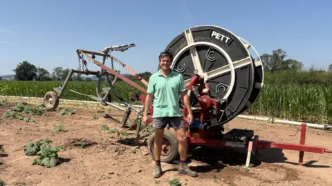 A man stands in his farmer's field in front of a large farming machine. He wears a light-green polo shirt and dark shorts.