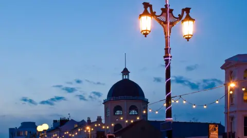 Worthing Pier at dusk. A street lamp is illuminated with a string of lights underneath. The sky is fading with some blue clouds scattered. The top of the pier is visible at the bottom of the photo. 