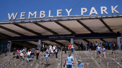 Football fans walk down stairs from Wembley Park station towards Wembley Stadium. It is clearly  sunny day. The fans are most wearing sky blue Coventry City football shirts.