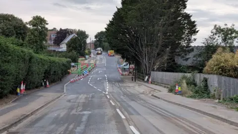 Single carriageway road with numerous cones along its edge and roadworks vehicles in the distance.