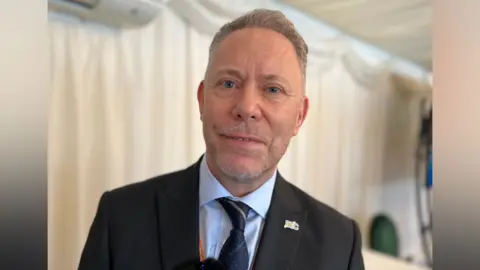 Michael Pemberton has short grey hair and blue eyes. He is wearing a dark blue tie and a black suit jacket with a BEC brooch. 