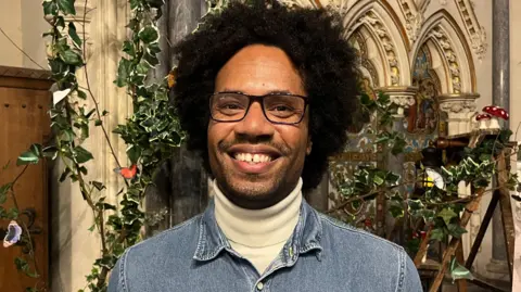 Choir member James Joseph pictured at the chapel. He is wearing a denim jacket and is smiling into the camera. There is holly draped over a trellis in the chapel behind him.