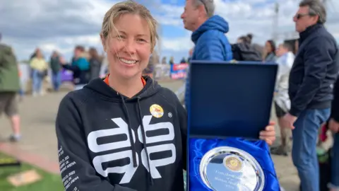 Lewis Adams/BBC Vicki Riley smiles as she holds up her medal. She wears a sweatshirt and around her are onlookers.