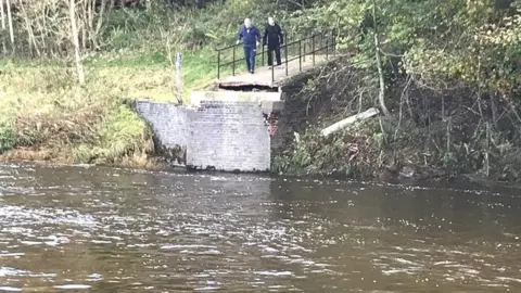 Two men standing at the end of a concrete path at the site of where a bridge used to span the river. The river, brown in colour, is shown with trees and greenery to the side