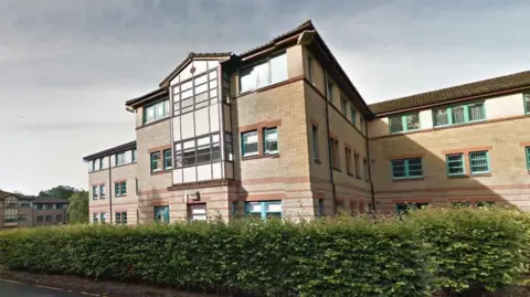 Google Alexander Court exterior from Google Maps - a three storey student residence building with tan bricks and brown brick accents, turquoise window frames. There are waist-height bushes around the building with the image taken from the car park at the rear.