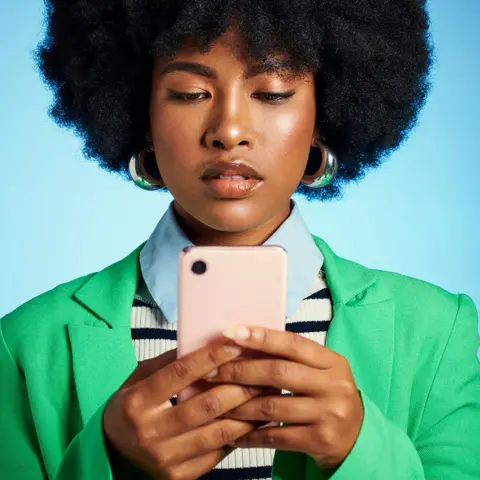 Getty Images A woman looking at her smartphone. she is wearing a green jacket, blue shirt and blue and white stripe top and is in front of a plain blue background