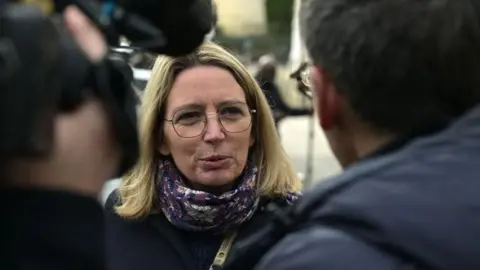 Getty Images A blonde-haired woman in glasses talks to reporters