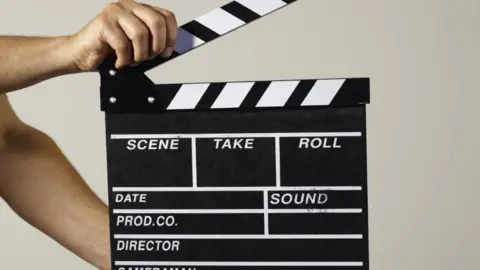 Getty Images A man is holding a black and white movie clapperboard against a cream-coloured wall