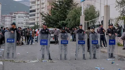 Getty Images A line of six riot police officers stand in a street in Turkey with clear shields with the word 'polis' on the front. There is a small crowd of people behind them and behind that stands several white and brown apartment buildings.
