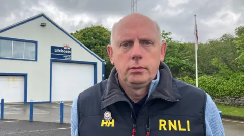 A man is wearing a blue fleece and black body warmer with the words: 'RNLI' on the front. He is also wearing a lapel pin. He is standing in front of a large building and a flagpole is seen behind him.