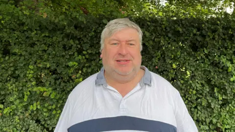 A middle-aged man wearing a grey shirt looks towards the camera with a hedge in the background.