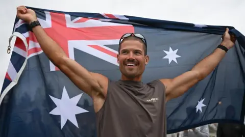 British ultra endurance runner William Goodge celebrates, holding an Australian flag, after crossing the finish line at Bondi Beach