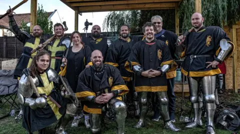 Knight Illuminations Group of people gathered outdoors under a wooden shelter, wearing partial medieval armour and black tunics with yellow trim. Some hold wooden training weapons or axes, while others rest hands on armour. Metal helmets and gear lie on the ground nearby. 