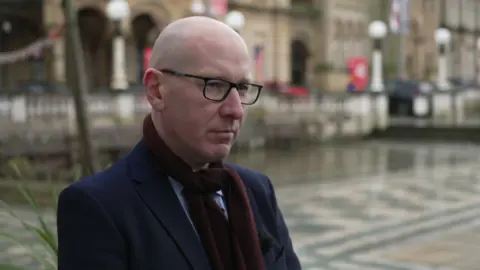 Patrick Hurley, being interviewed by BBC North West Tonight, is wearing a dark blue jacket, a light blue shirt and a burgundy scarf. He is largely bald with the rest shaved, and is wearing glasses. A blurred-out street is behind him.