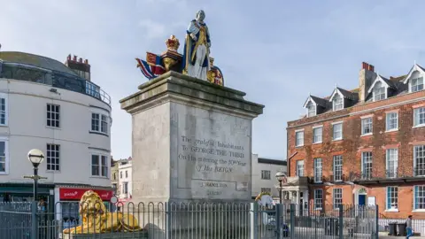 Ian Capper/Geograph Looking up at the King George III statue on top of a large stone plinth. The painted statue depicts the king standing between a crown and a pile of books. At the base of the plinth is a gold lion and a white and gold unicorn.