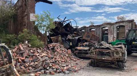 A pile of red bricks next to a derelict building and twisted metal at the Salthouse Mills site in Barrow. There are bushes growing out of the derelict red-brick building. There is a heavy plant machine in the background, in front of part of another derelict building.