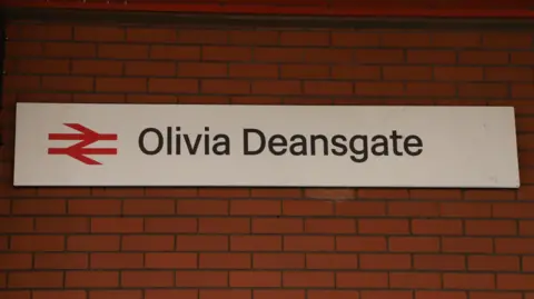 A train station sign reading 'Olivia Deansgate' hangs on a red brick station wall
