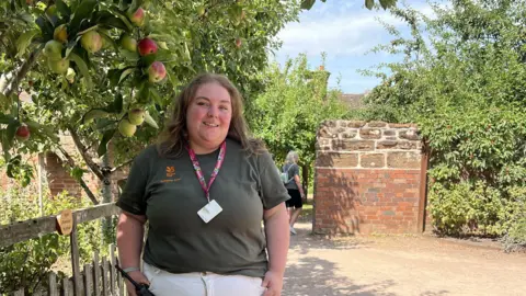A woman with long light brown hair is wearing a green/brown t-shirt and white trousers. she is standing next to a wooden fence with an apple tree hanging over it