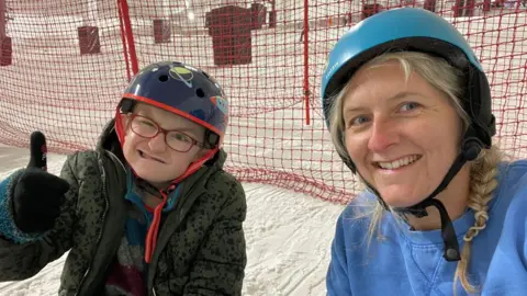 Hugo and Claire Reece on a ski slope. He is on the left and wearing glasses, a black helmet and dark fleece and has his right thumb up while smiling. She is on the left wearing a light blue helmet over plaited blonde hair and a blue sweatshirt and also smiling. They are sitting on snow and behind them is a red net. 