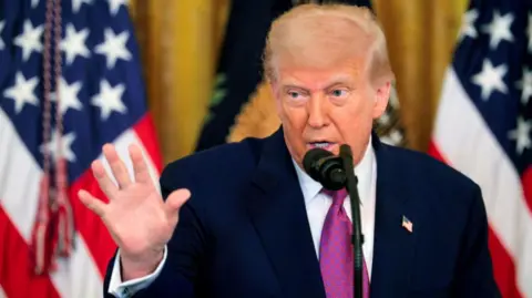 Donald Trump in a suit with his hand raised, standing in front of US flags