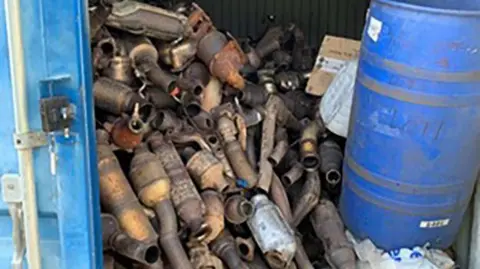 A blue shipping container filled with rusted metal car exhaust parts. The catalytic converters are stacked up next to a blue barrel.