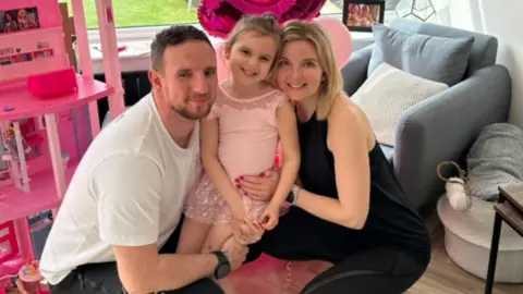 Victoria Skinner Tor, with her husband, Joe and daughter Isla Rose, both parents are crouching down, holding their daughter, wearing a pink dress, in a room, with pink toys and furniture. They are all looking at the camera. 