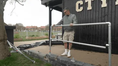 BBC/Jasmine Lowe Tony Henderson is standing holding a metal railing attached to stone slabs outside of the youth hub. He is staring down at another metal railing that has been vandalised. 