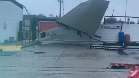 A blurry photo of the front of Sawbridgeworth Town's club, with the roof blown off and resting on a broken wall. There are red barriers lyig on the floor, and you can see half a sign with the words "TOWN FC". The sky is very grey.