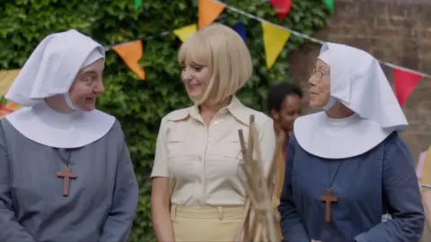 Three actors stand in a row in a garden - two dressed as nuns on either side of a woman in a yellow shirt with a blonde bob
