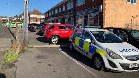 Two police cars are parked up outside a parade of shops with flats above them. There are other cars also parked up along the street, with houses visible in the background. 